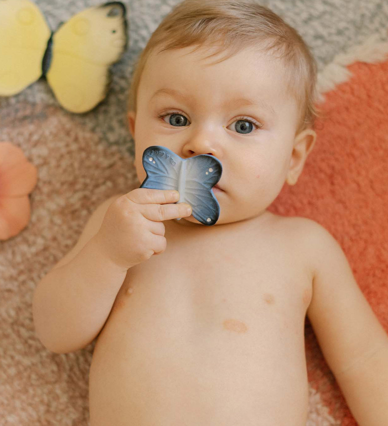 A child holding the Oli & Carol Natural Rubber Mini Teether - Blues The Butterfly to their mouth, and laying on a colourful rug