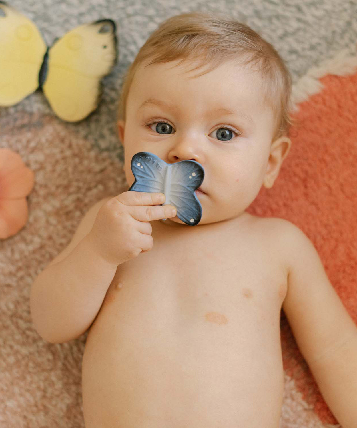 A child holding the Oli & Carol Natural Rubber Mini Teether - Blues The Butterfly to their mouth, and laying on a colourful rug