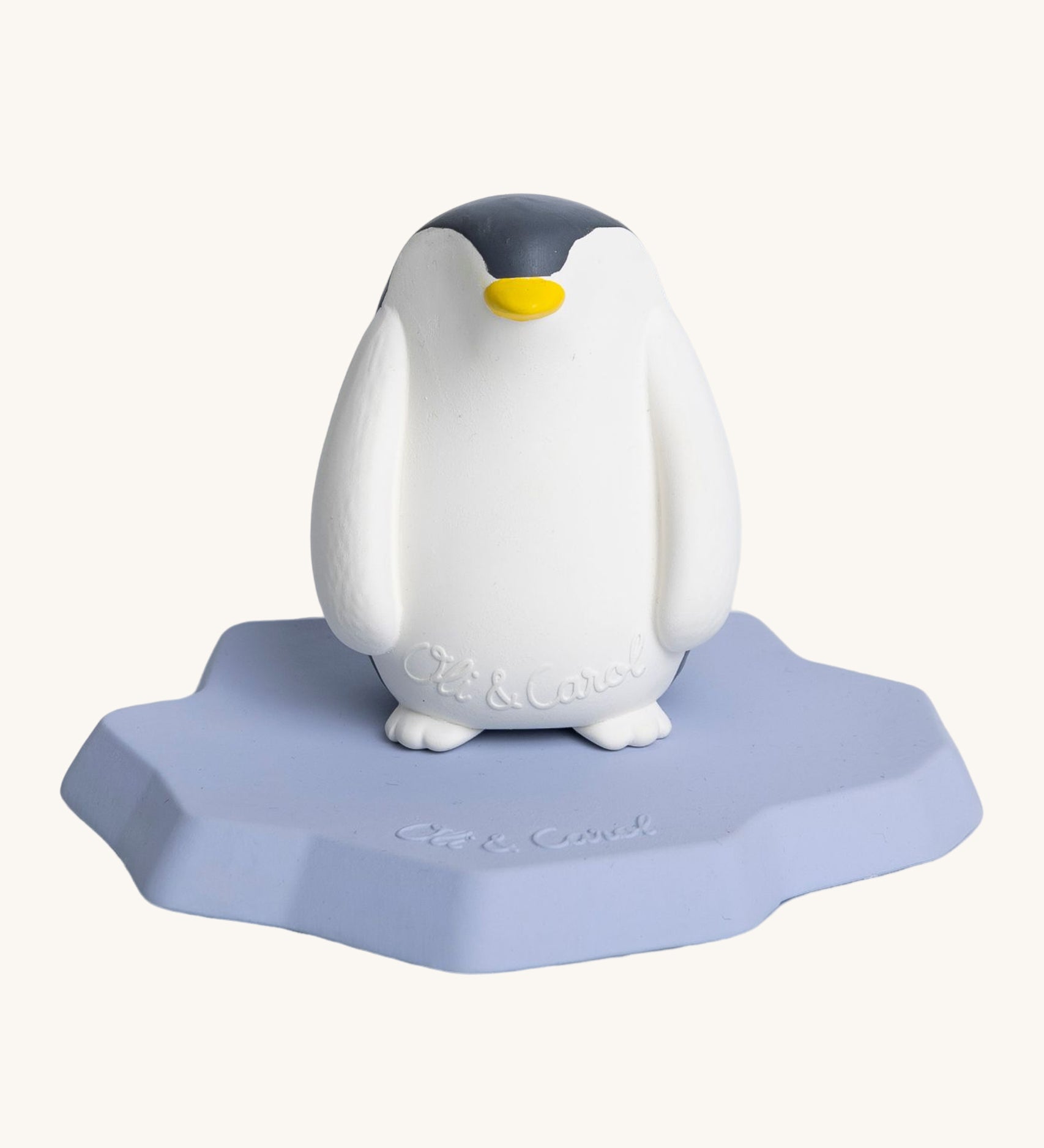 Oli & Carol Penguin Bath Toy. A bright white penguin, natural rubber bath toy is sitting on a flat, light blue/lilac ice berg on a cream background. The penguin is white and black, has a slightly textured body and has a yellow beak. 