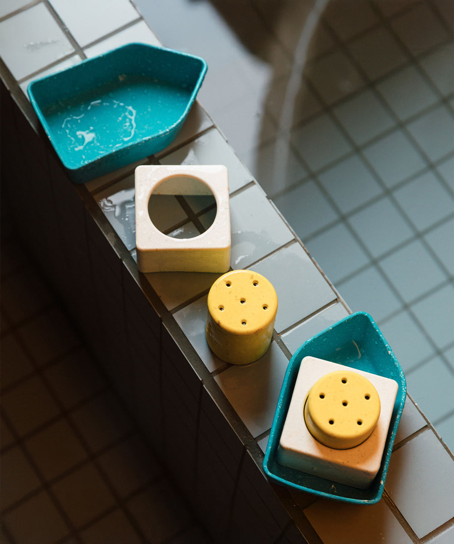 Oli & Carol Boat Bath Toy - Yellow pieces all lined up in a row on the side of a light blue tiled bath