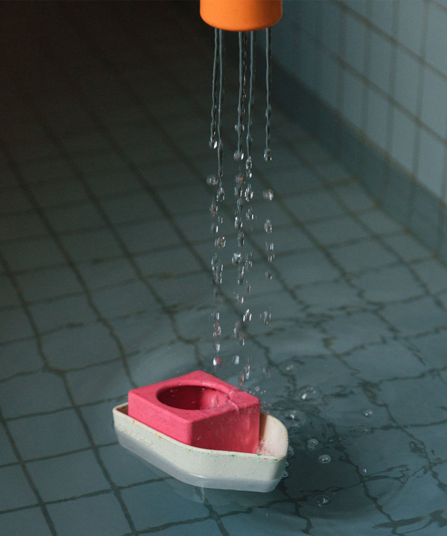 The orange part of the Oli & Carol Boat Bath Toy - Orange, sprinkling water over the rest of the boat parts. The boat is floating on water