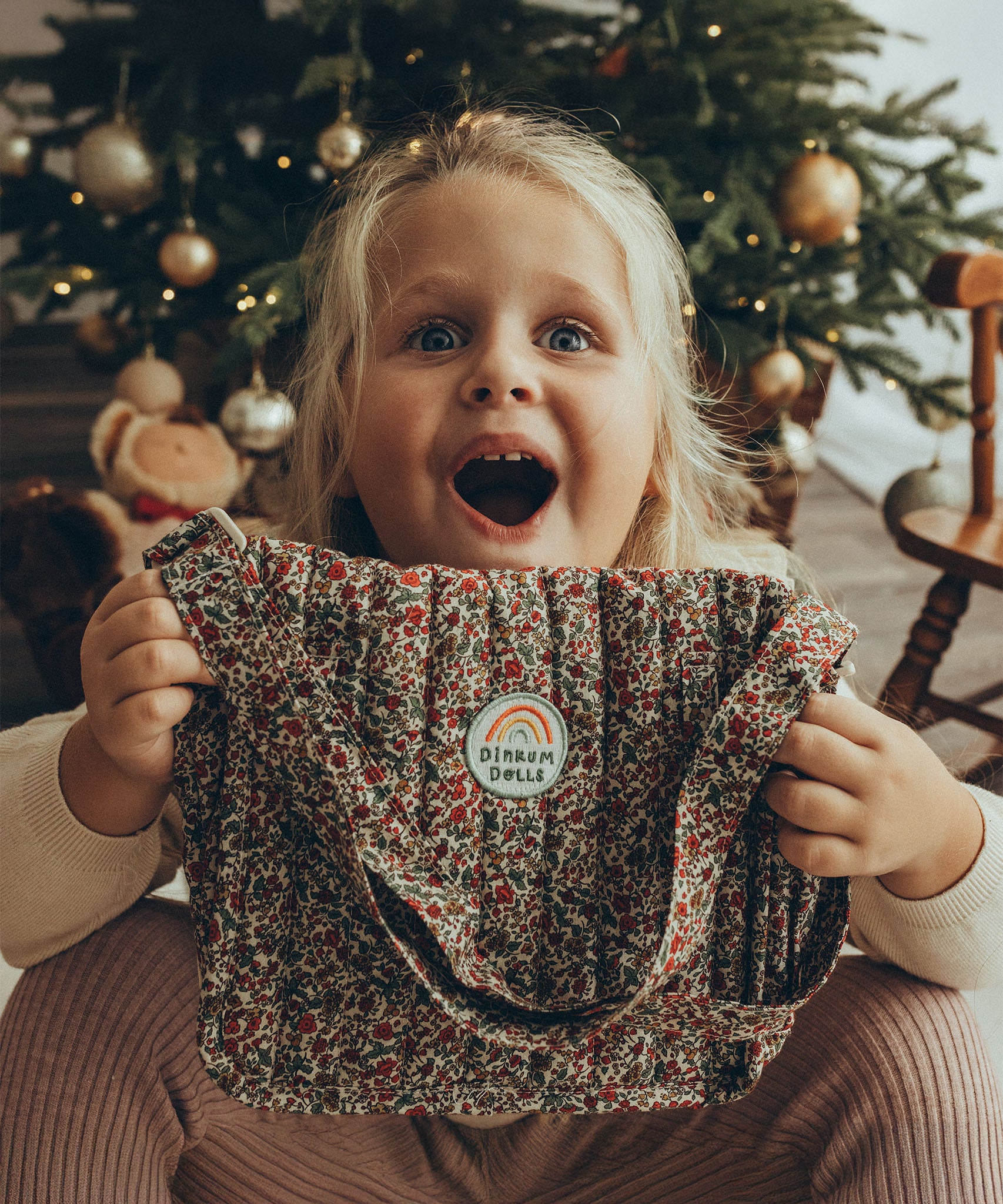 A child looking very excited to have received the Olli Ella Carrie Convertible Changing Set - Garland. The child is holding the toy changing bag in their hands, with a surprised and happy expression on their face.