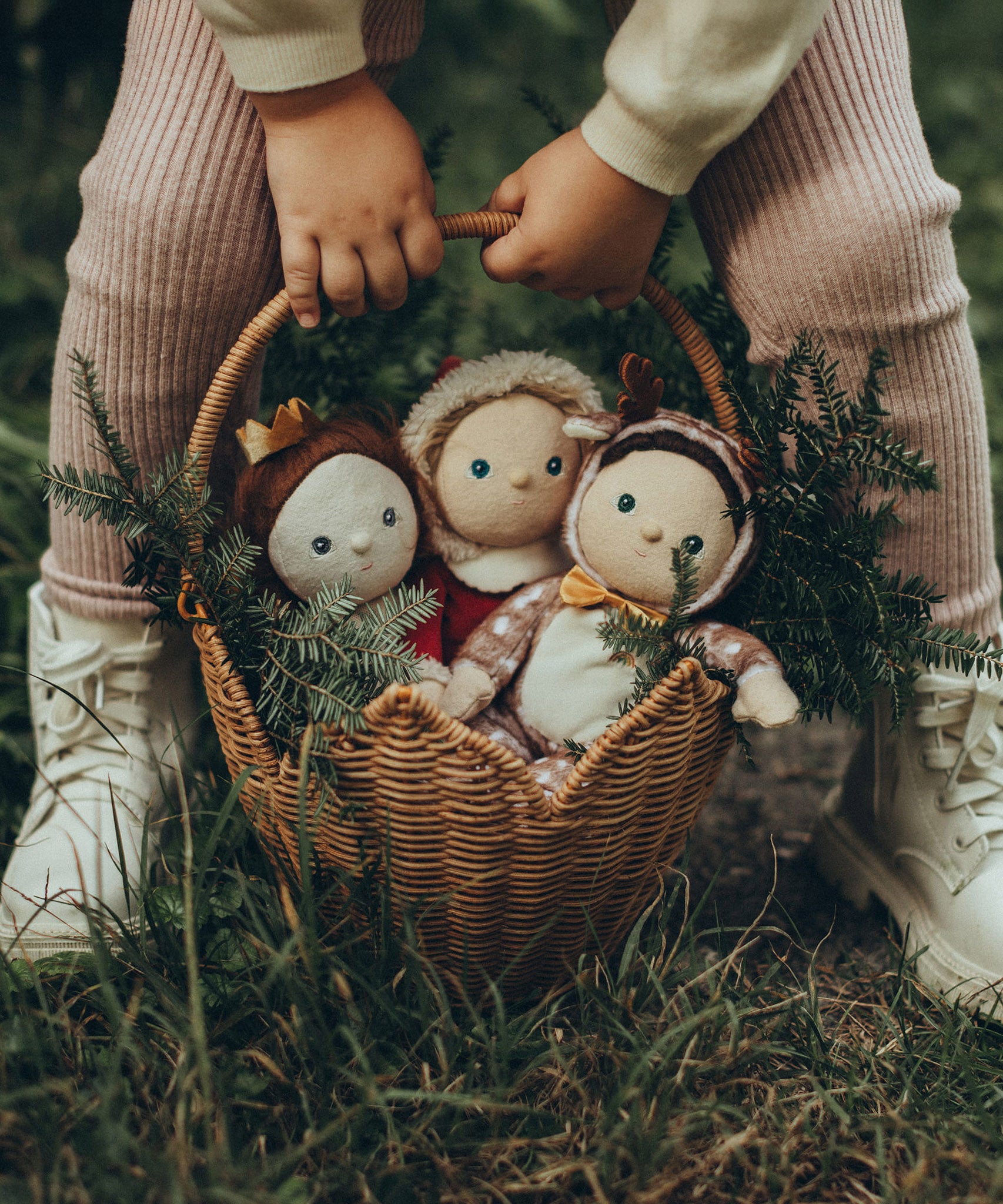 A child is holding the Olli Ella Rattan Poinsettia basket by the handle, in their hands. The basket is on a grassy floor with Christmas Dinky Dinkums inside, and small pine tree branches to give a festive feel.