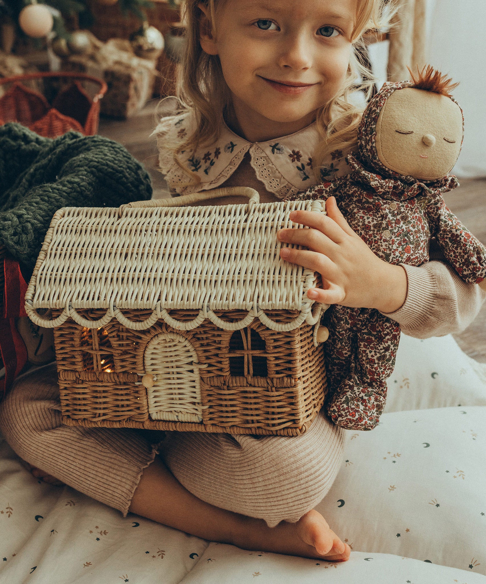 A child is holding the Olli Ella Rattan Gingerbread House Basket - Natural & Straw, which has some fairy lights inside. This decorative, eco-friendly basket is perfect for storing small toys, and great for gifting.