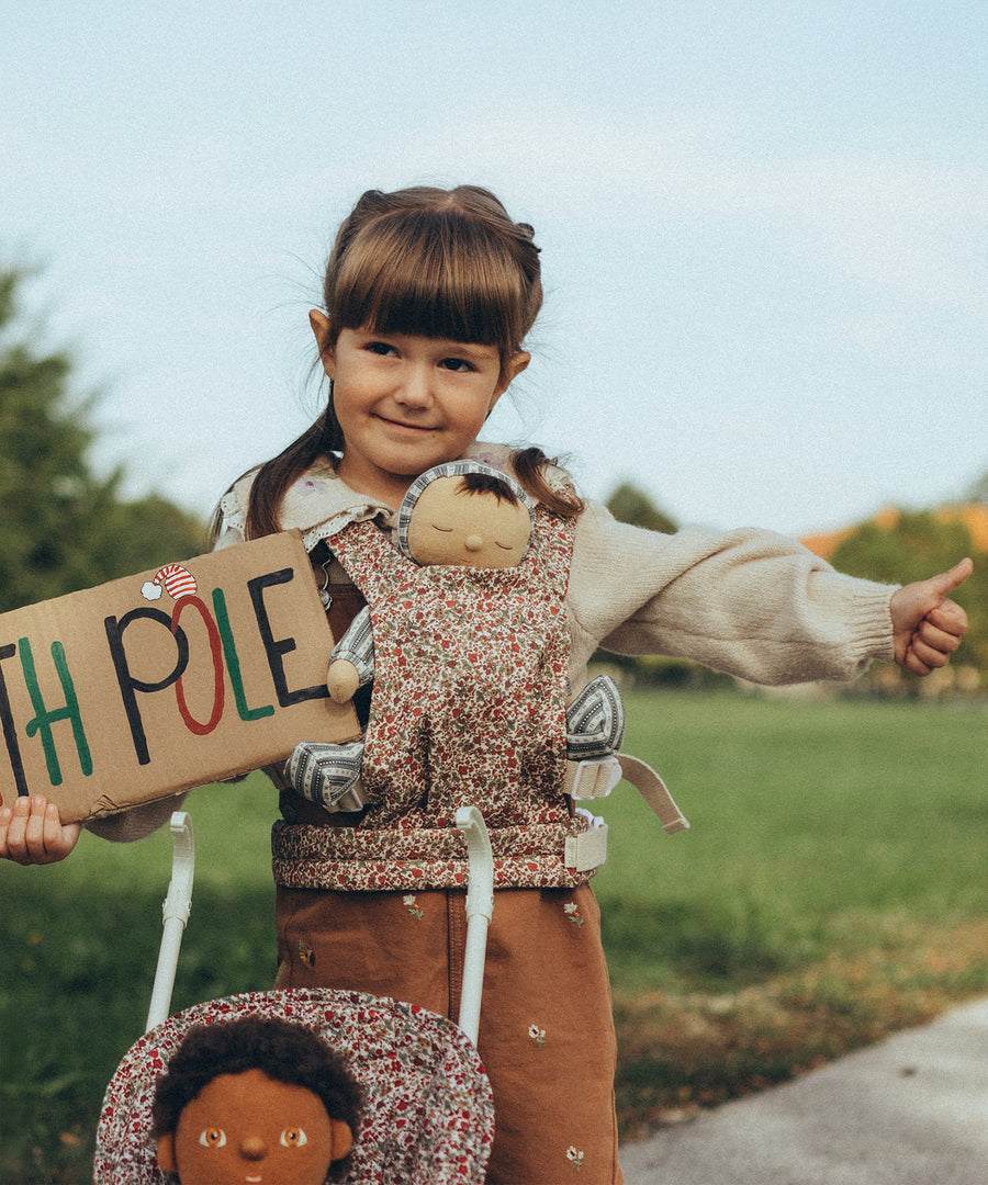 A child holding a cardboard sign saying "North Pole" in colourful letters whilst wearing an Ollie Ella Dinkum Doll in the Olli Ella Dinkum Dolls Carrier - Garland. There is also the top part of an Olli Ella Stroller by the childs legs, with a Dinkum Doll sat inside.