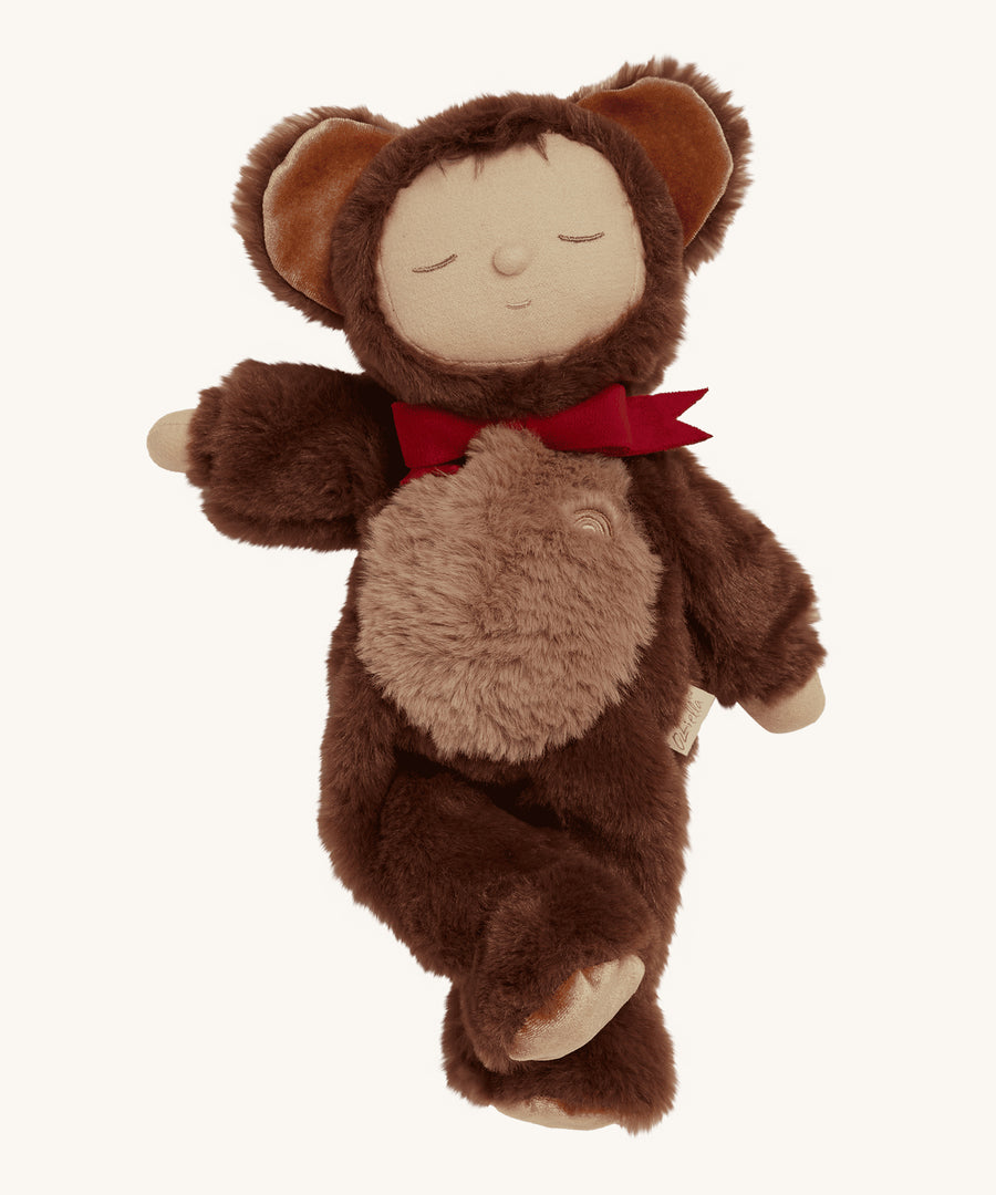 Olli Ella Cozy Dinkums - Teddy Twiggy is a soft body doll, with a soft and fluffy chocolate brown outfit which has a hood and teddy ears on top, and a red bow tie. The image is on a cream background.