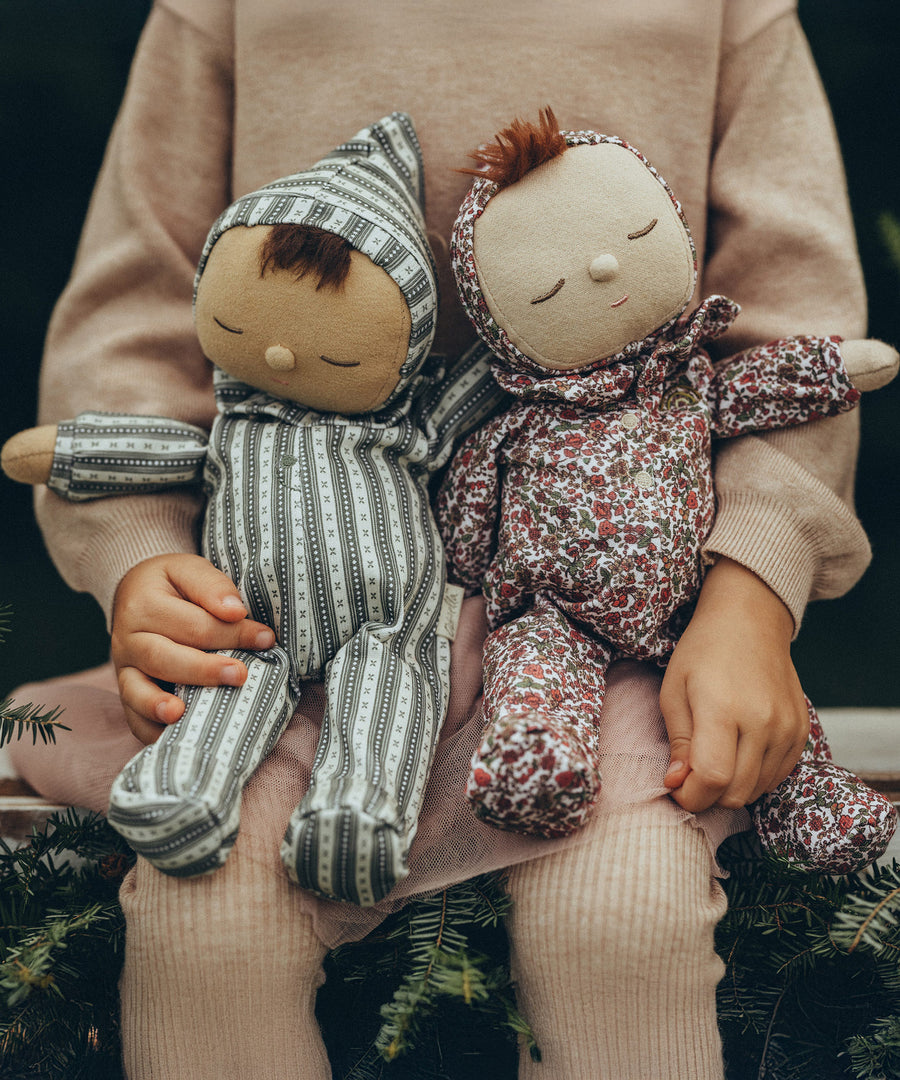 Olli Ella Dozy Dinkum Doll Pudding and Pie, sat on a child's knee. Pie is on the left and wears a vintage green and white stripe outfit with a hood. Pudding is on the right and wears a vintage style, floral outfit with a hood.