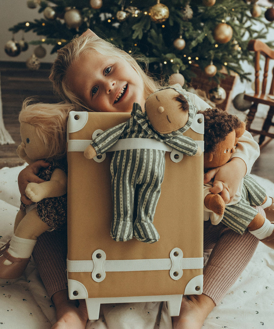 A child happily playing with their Olli Ella Dolls at home, with a Christmas tree in the background. Olli Ella Dozy Dinkum Doll Pie - Clover is attached to the back of an Olli Ella See-Ya Suitcase in front of the child, ready for a journey. The child also holds two other Olli Ella Dinkum Dolls in each arm