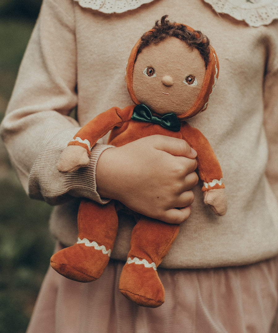 A child is holding Olli Ella Georgie Gingerbread in their hand. The child is holding the doll close to their torso