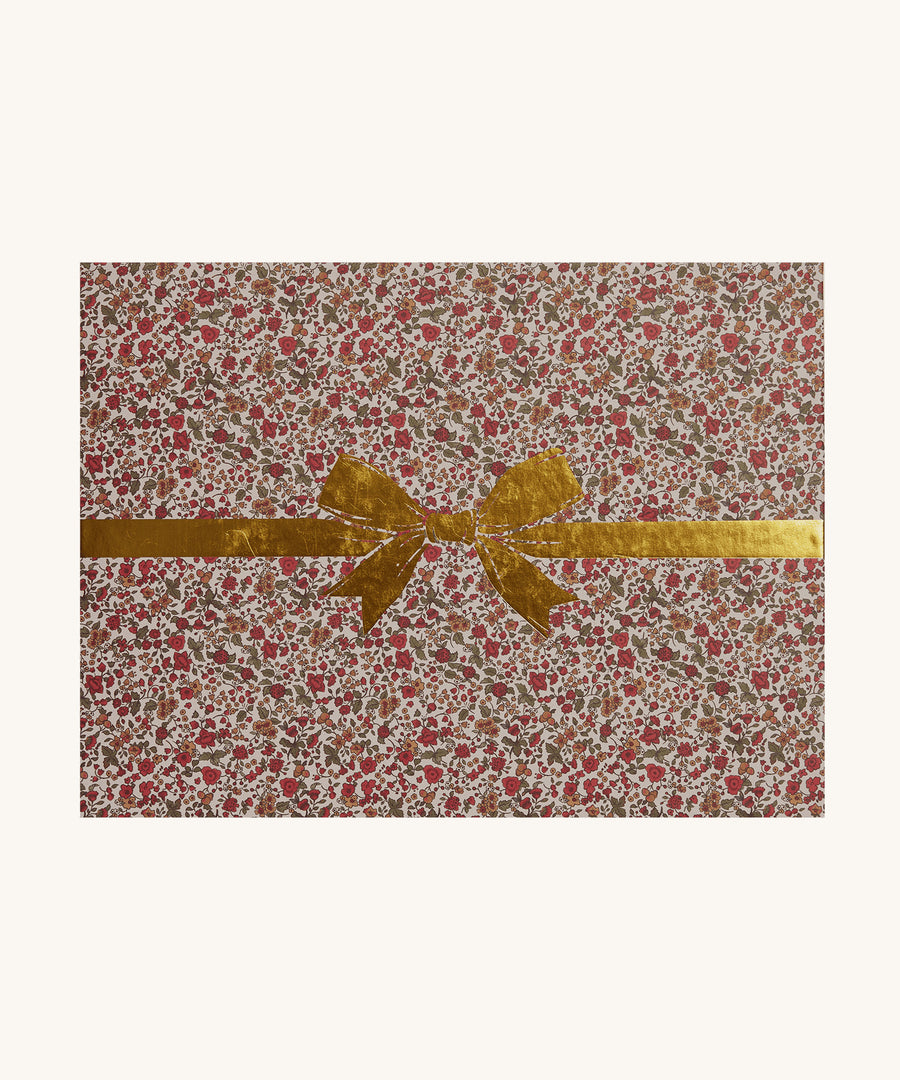 The packaging for the Olli Ella Dinkum Dolls Carrier in Garland. The packaging has the same colourful, vintage, floral print and has a gold painted bow on top. The image is on a cream background