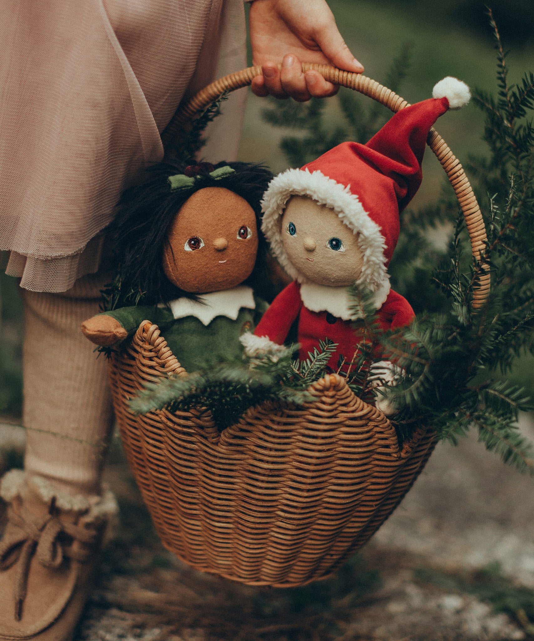 A child is carrying their Festive Jolly Dolly Dinkum Dolls, Paige Pudding and Sammie Santa, in a natural Ollie Ella Poinsettia Carry Basket. Inside the basket with the dolls are branches from a pine tree, to give a festive feel.