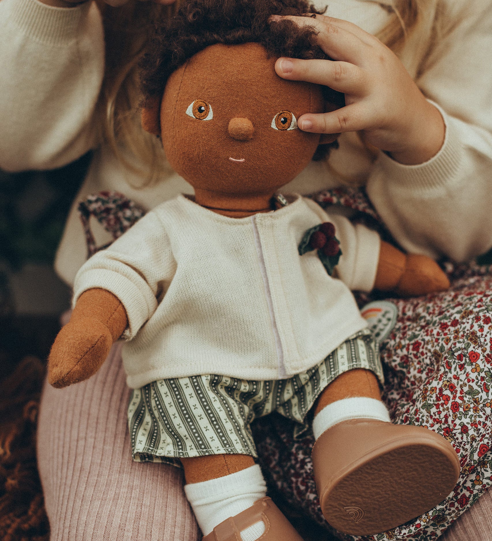 A child sat with an OllI Ella Dinkum Doll on their knee, which is wearing the Olli Ella Dinkum Doll Holiday Outfit Set.