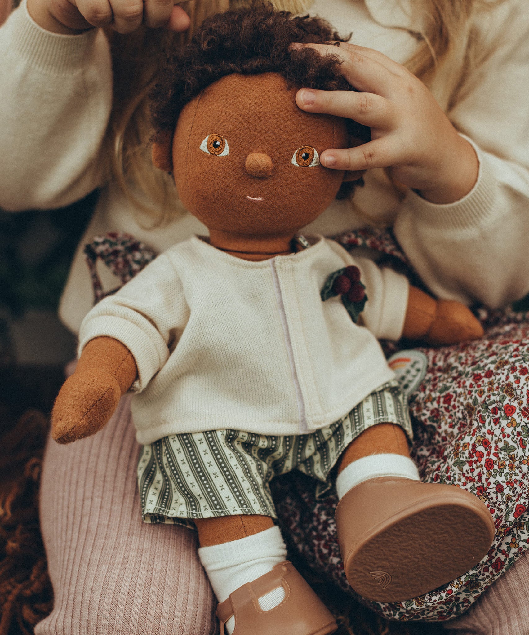 A child sat with an OllI Ella Dinkum Doll on their knee, which is wearing the Olli Ella Dinkum Doll Holiday Outfit Set.