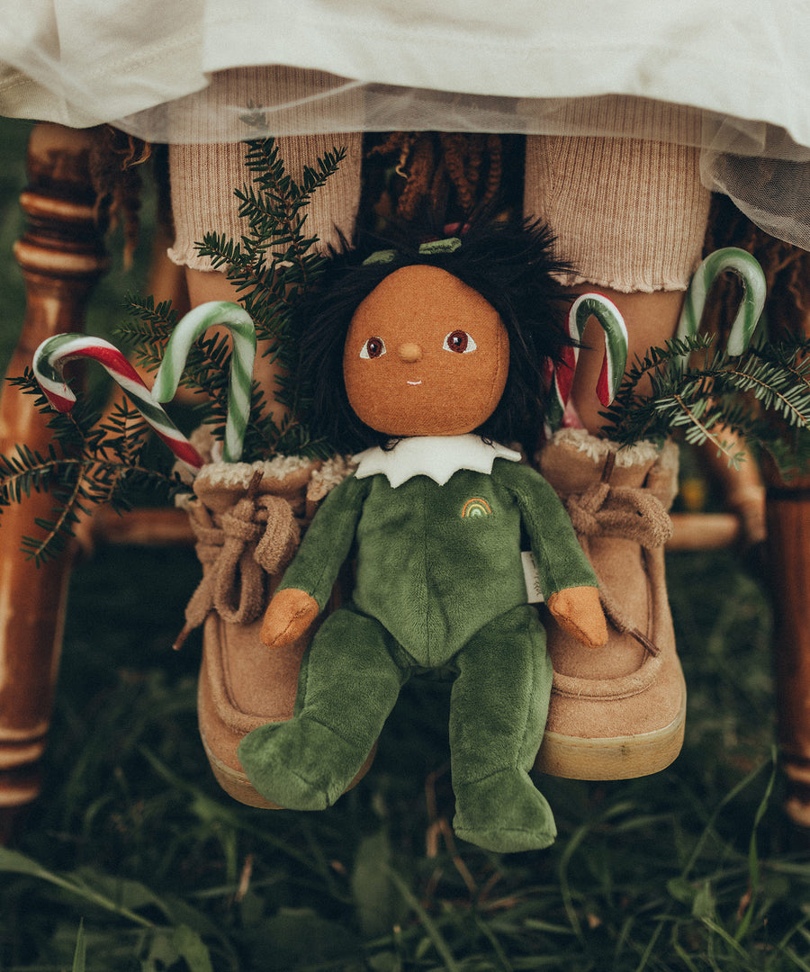 Olli Ella Dinky Dinkum Doll - Paige Pudding is sat between a child's feet. The child is wearing tan/pink boots with red and green candy canes and small pine tree branches poking out to give a festive feel. 