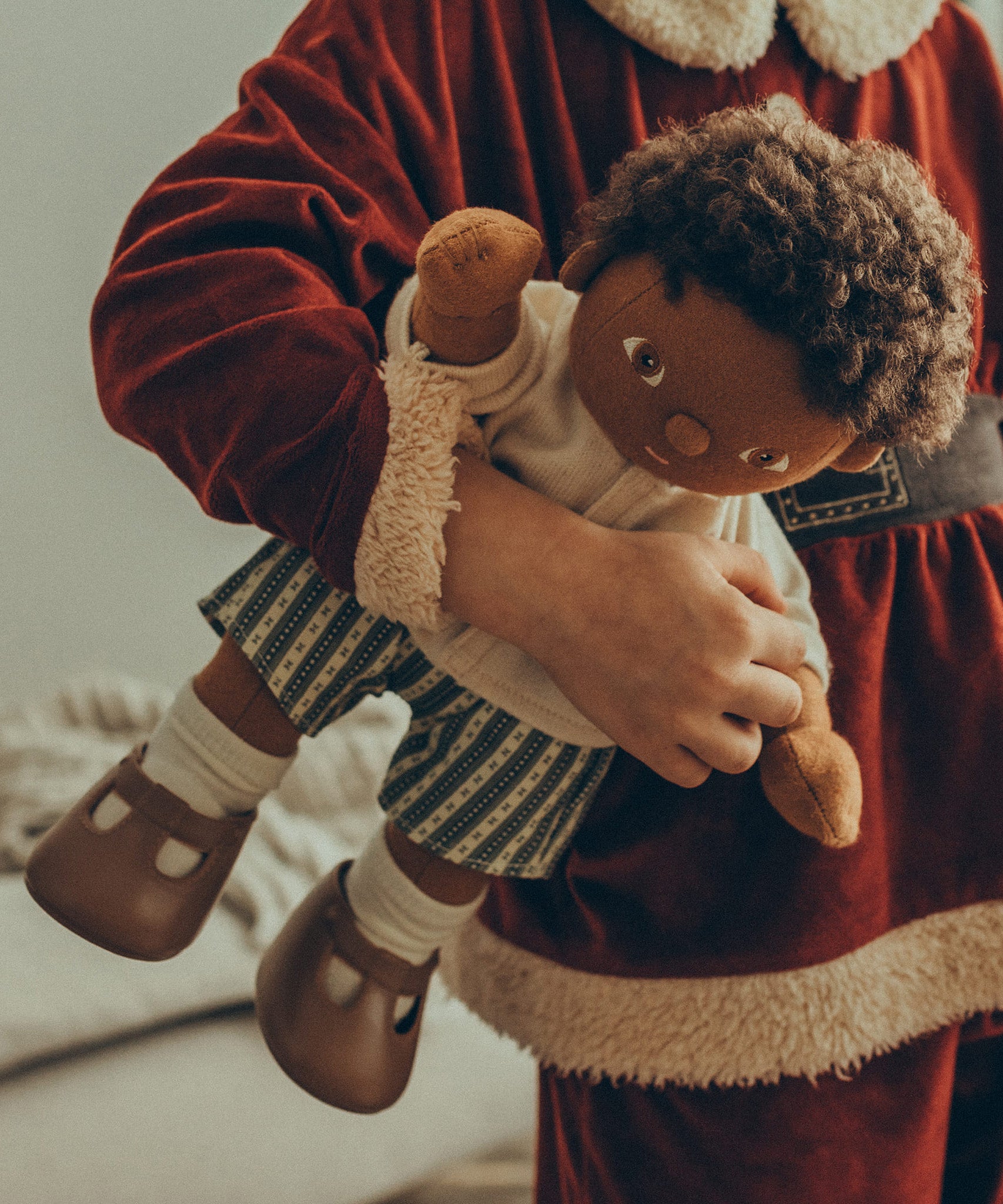 A child wearing a Santa outfit, is holding a Olli Ella Dinkum Doll under their arm, which is wearing the Olli Ella Dinkum Doll Holiday Outfit Set