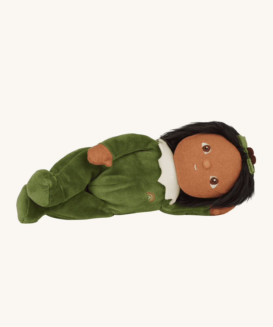 Olli Ella Dinky Dinkum Doll - Paige Pudding is lying on their side. Paige wears a soft, green velvet onsie with a white icing collar, dark hair, brown skin and an adorable holly berry and leaf in its hair. The image is on a cream background