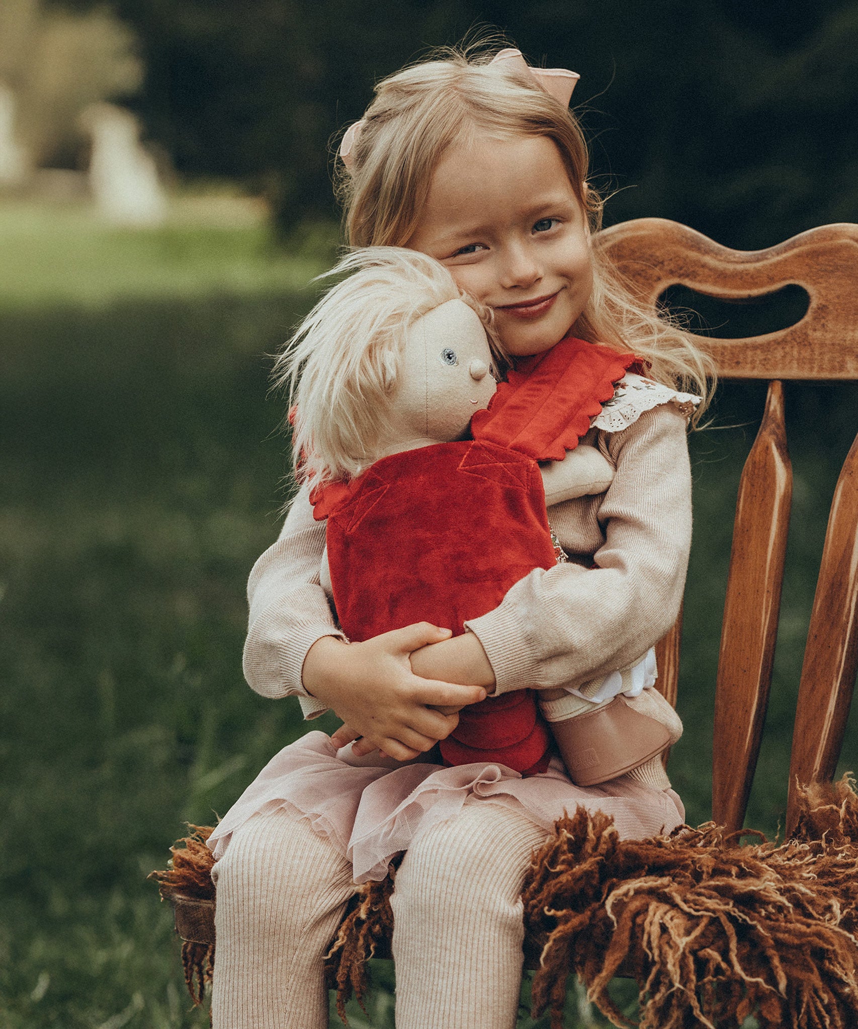 A child is sat down on a antique wooden chair, and is carrying a Dinkum Doll in the Olli Ella Dinkum Dolls Petal Carrier - Red Velvet. The child is happy and is cuddling the doll. The chair has a brown fluffy rug on the seat.