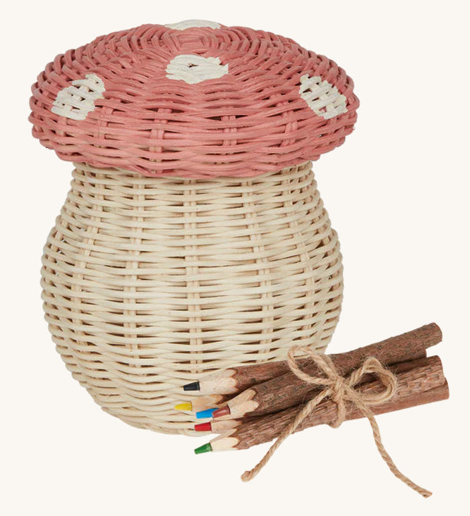 An Olli Ella Porcini mushroom shaped Basket with a pink spotted lid, and wooden twig colouring pencils wrapped together with string next to it pictured on a cream background