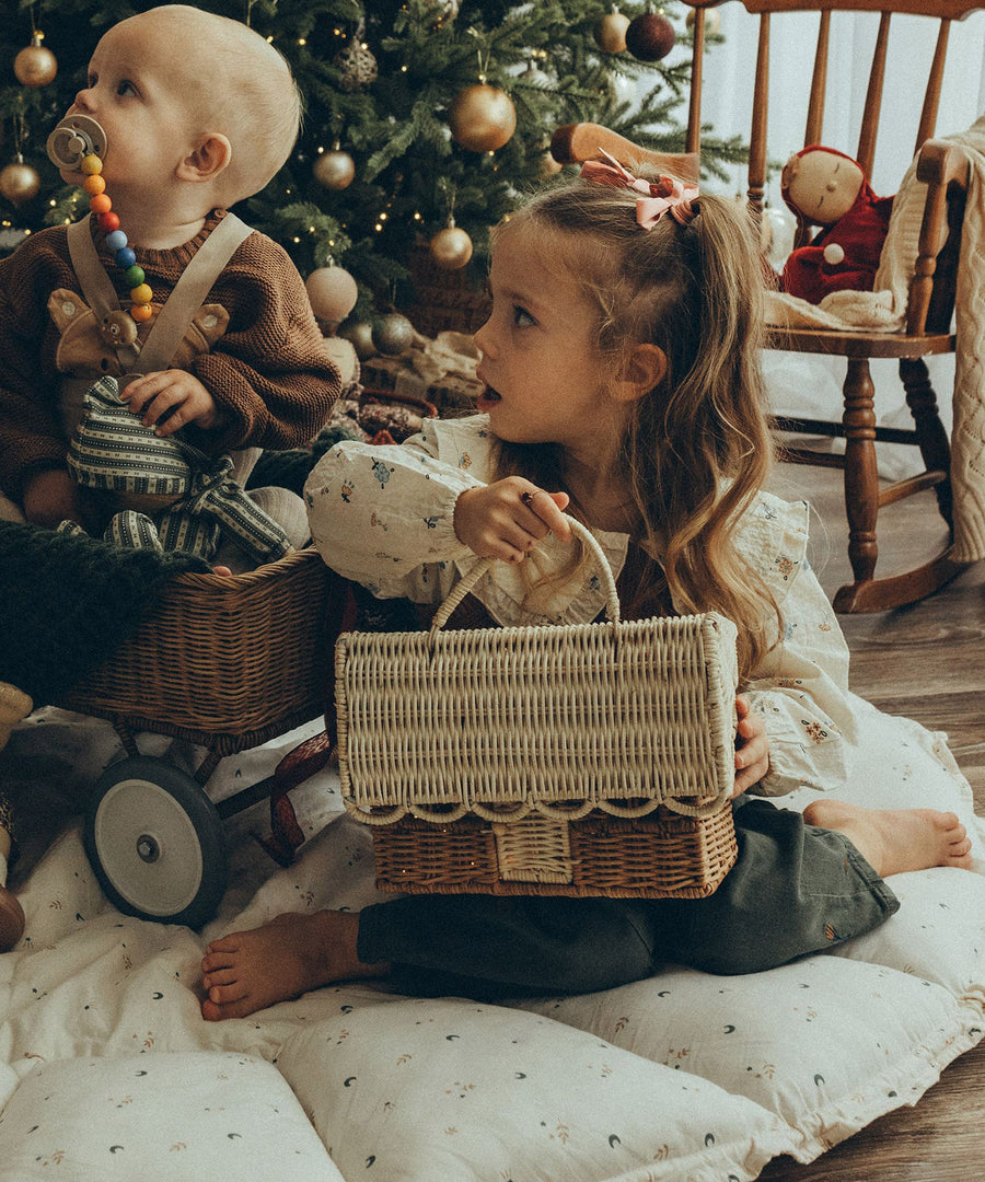 A child holding the Olli Ella Rattan Gingerbread House Basket whilst sat on a white cushion. A Christmas tree is in the background and a child is sat behind playing with an Olli Ella doll