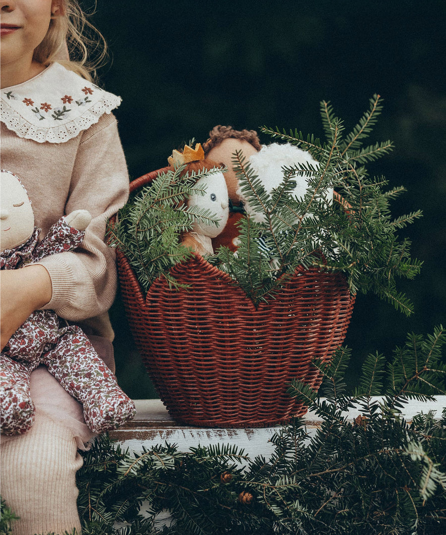 Olli Ella Christmas Dinky Dinkum dolls are in the red Poinsettia Carry Basket, and are hiding behind pine tree branches. A child is sat next to the basket which is on a white wall, and is holding a Dozy Dinkum doll in their hands