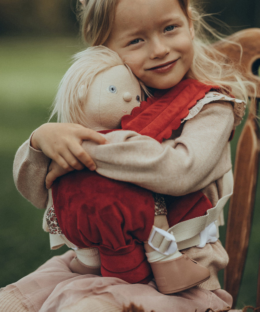 A child is sat down on a antique wooden chair, and is carrying a Dinkum Doll in the Olli Ella Dinkum Dolls Petal Carrier - Red Velvet. The child is happy and is cuddling the doll.