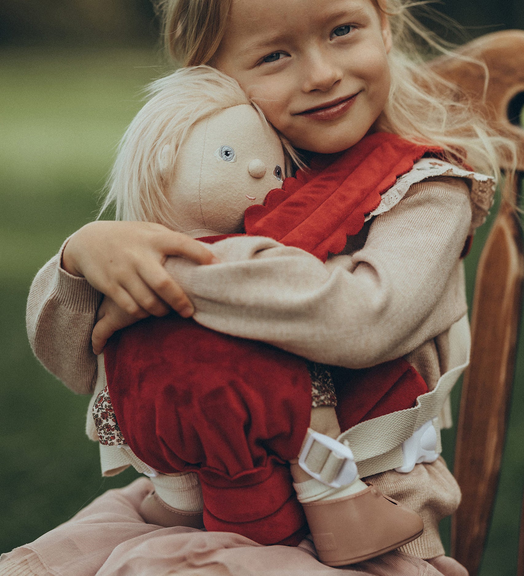 A child is sat down on a antique wooden chair, and is carrying a Dinkum Doll in the Olli Ella Dinkum Dolls Petal Carrier - Red Velvet. The child is happy and is cuddling the doll.