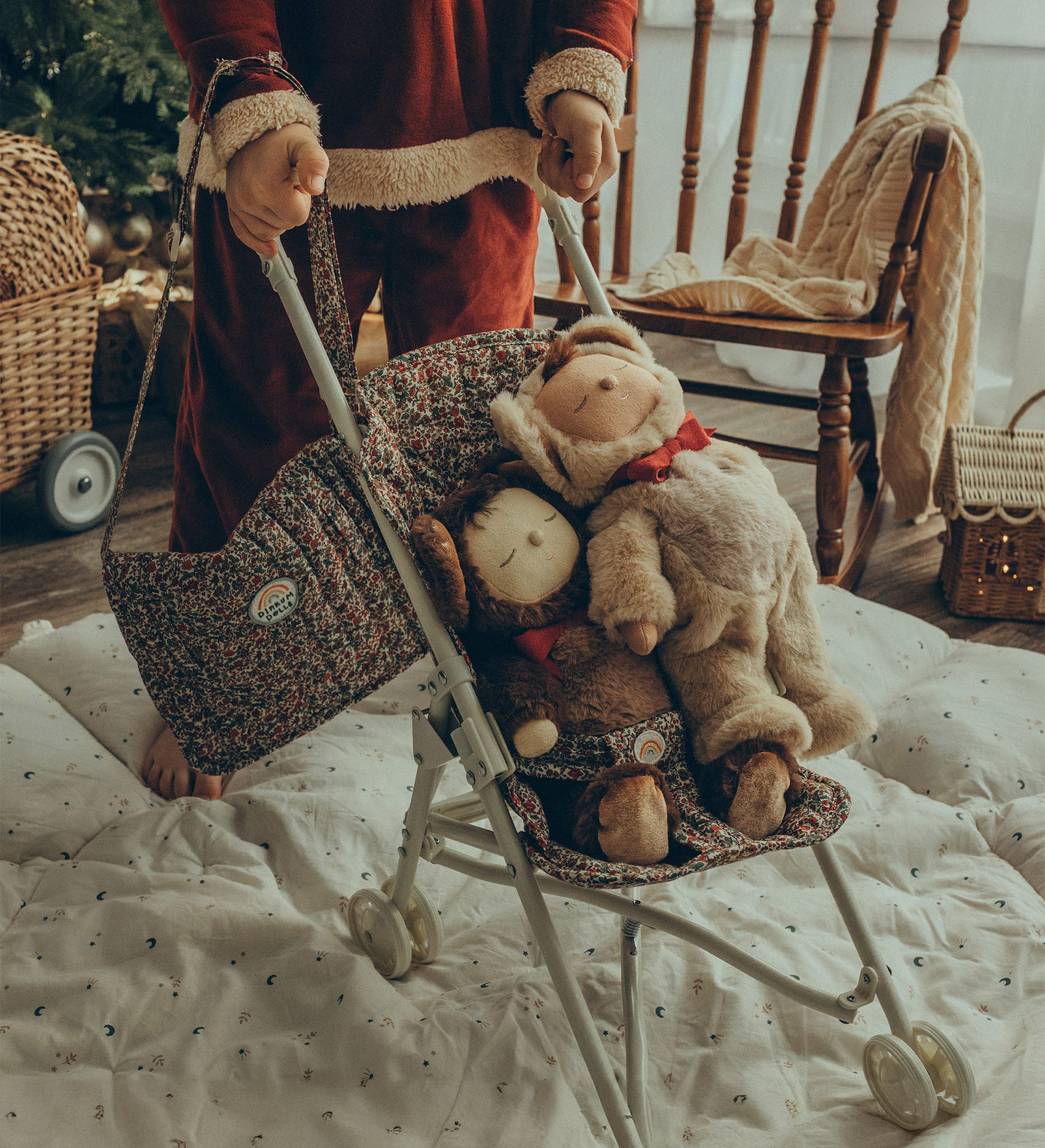 A child pushing two Olli Ella Cosy Dinkum Dolls in an Olli Ella Stroller. The child has the Garland Changing Bag hanging from their wrist, which can also be adjusted to fit over the shoulder.