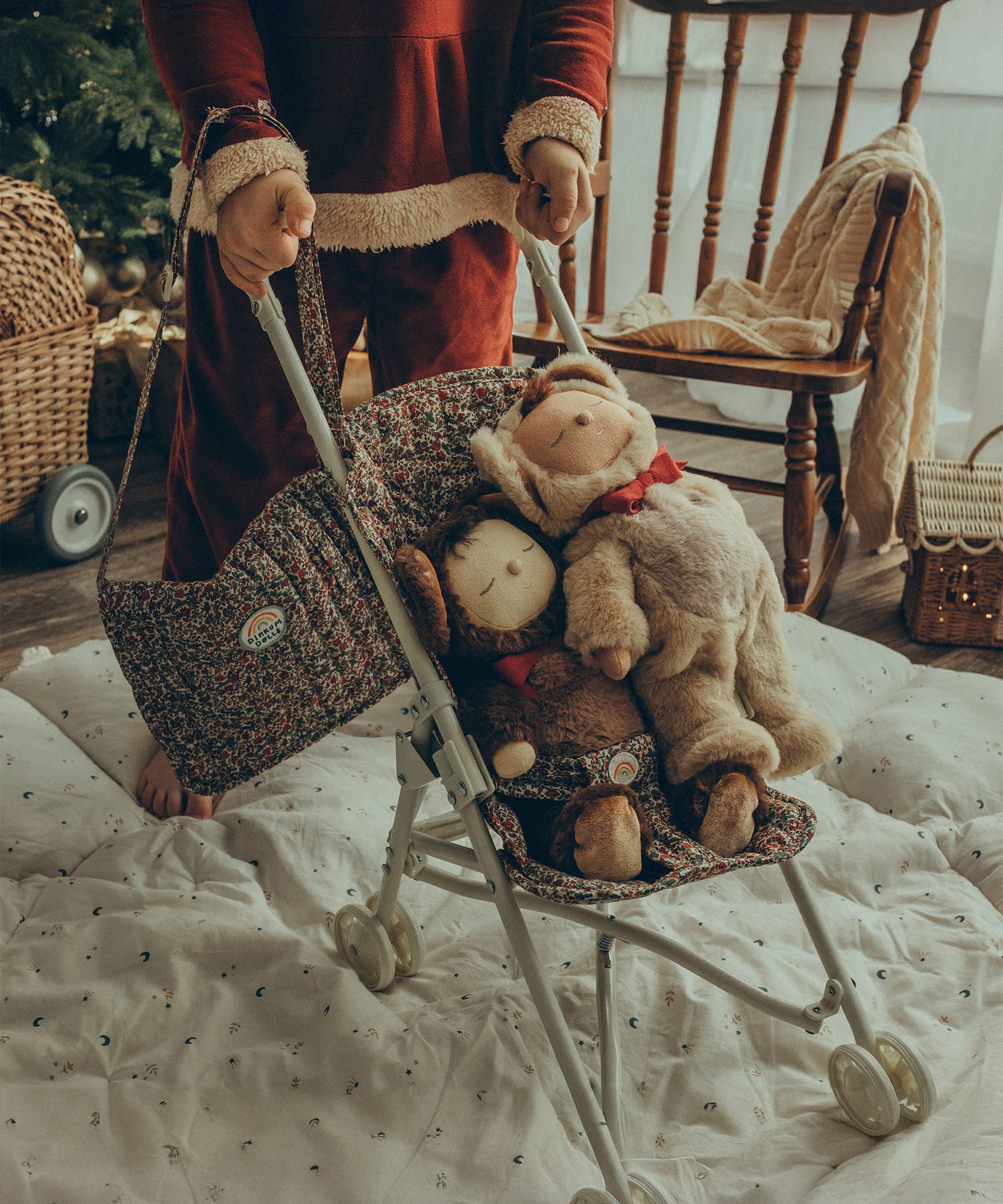 A child pushing two Olli Ella Cosy Dinkum Dolls in an Olli Ella Stroller. The child has the Garland Changing Bag hanging from their wrist, which can also be adjusted to fit over the shoulder.