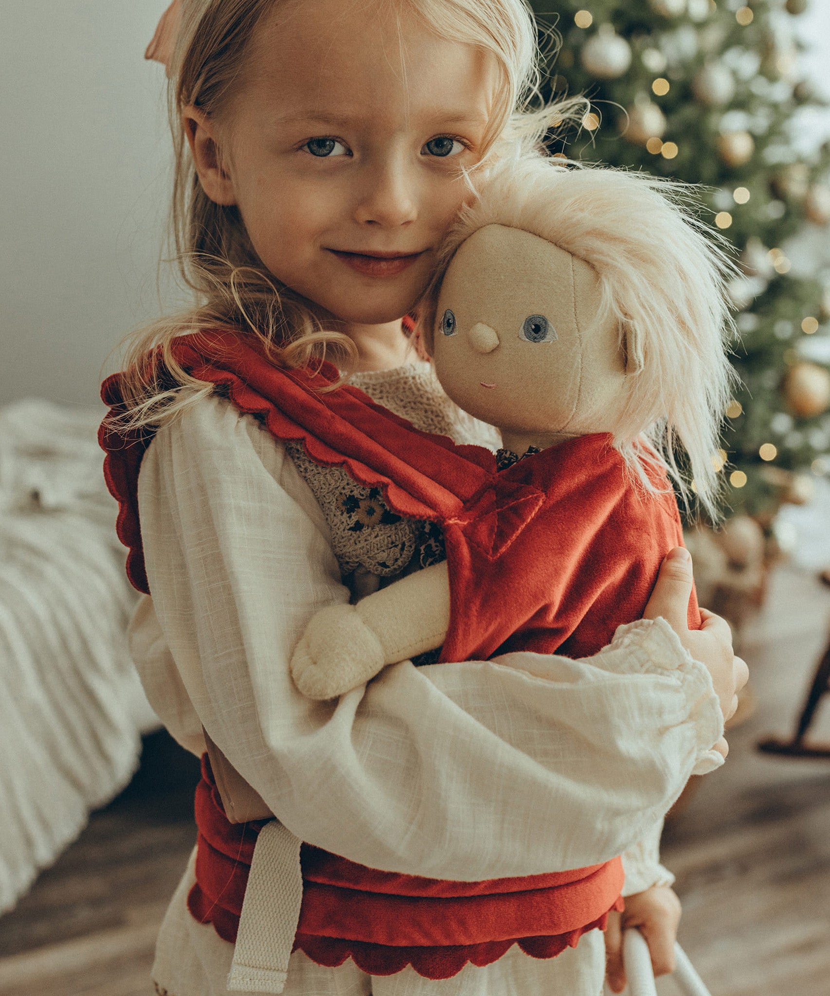 A child is carrying a Dinkum Doll in the Olli Ella Dinkum Dolls Petal Carrier - Red Velvet. The child is happy and is cuddling the doll, and there is a Christmas tree in the background