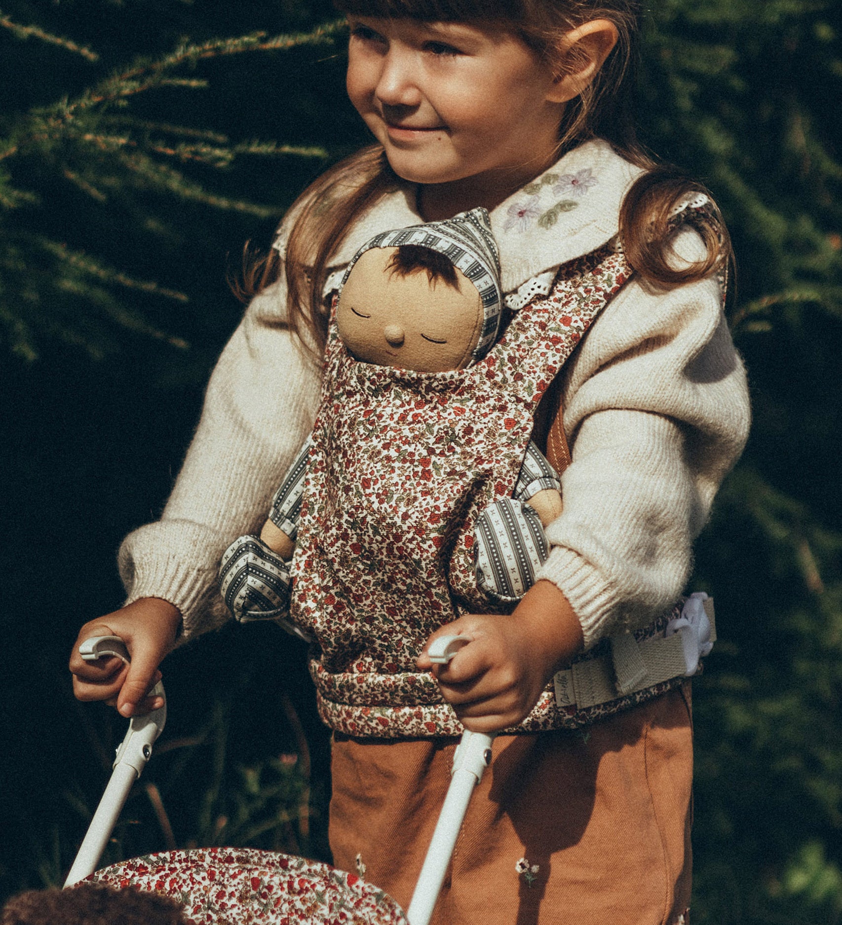 A child carrying a Dozy Dinkum Doll in a Olli Ella Dinkum Dolls Carrier in Garland print, on their front and is pushing a doll in an Olli Ella Stroller