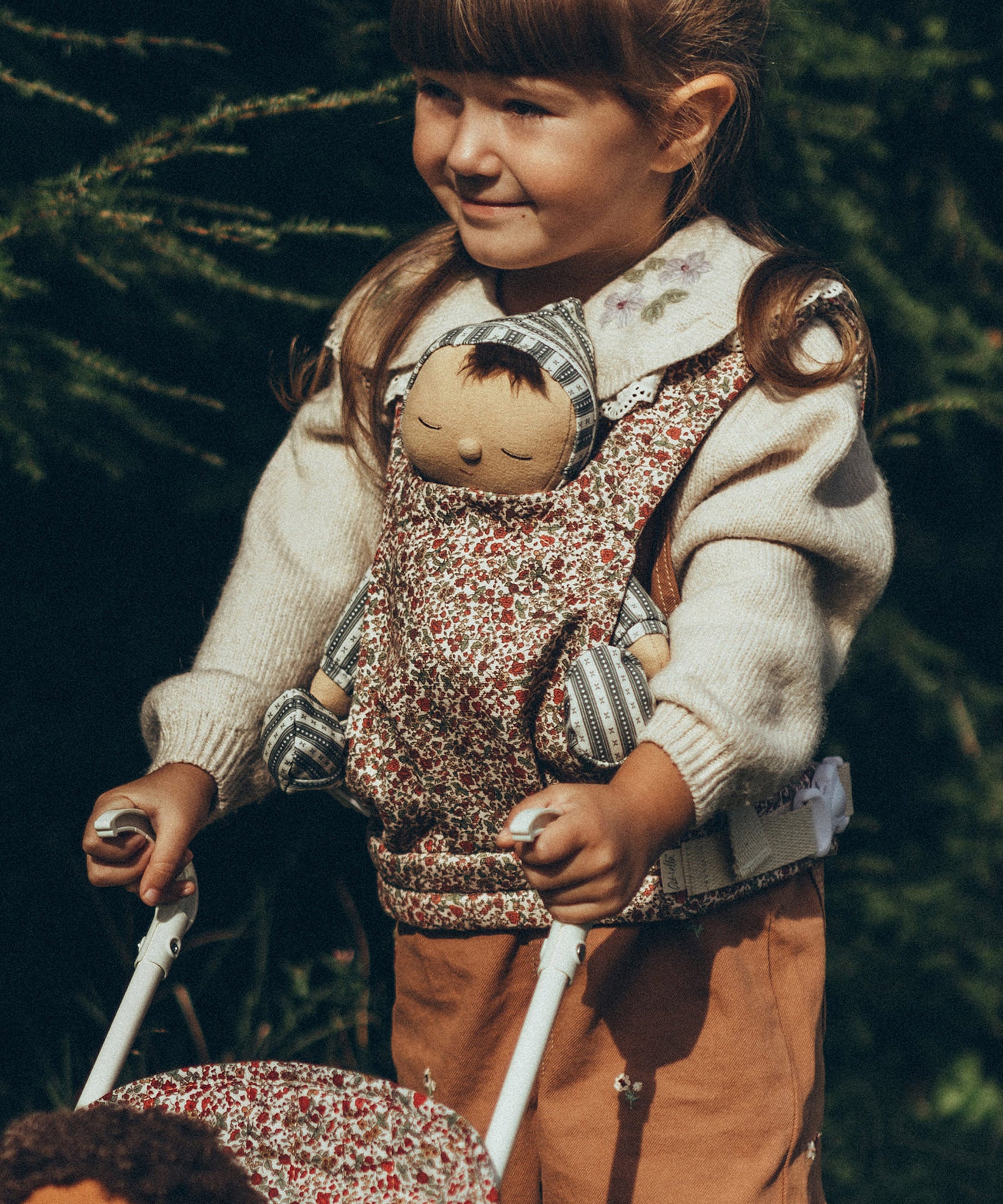 A child carrying a Dozy Dinkum Doll in a Olli Ella Dinkum Dolls Carrier in Garland print, on their front and is pushing a doll in an Olli Ella Stroller