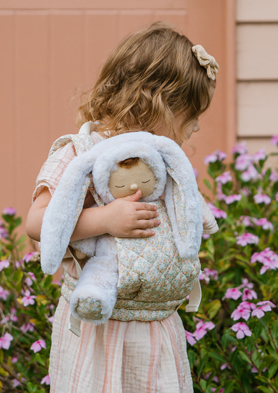 Olli Ella Cozy Dinkum Bunny Lola, soft toy with bunny ears being carried in a baby carrier by a child.  Outdoors spring image with flowers in the background. 