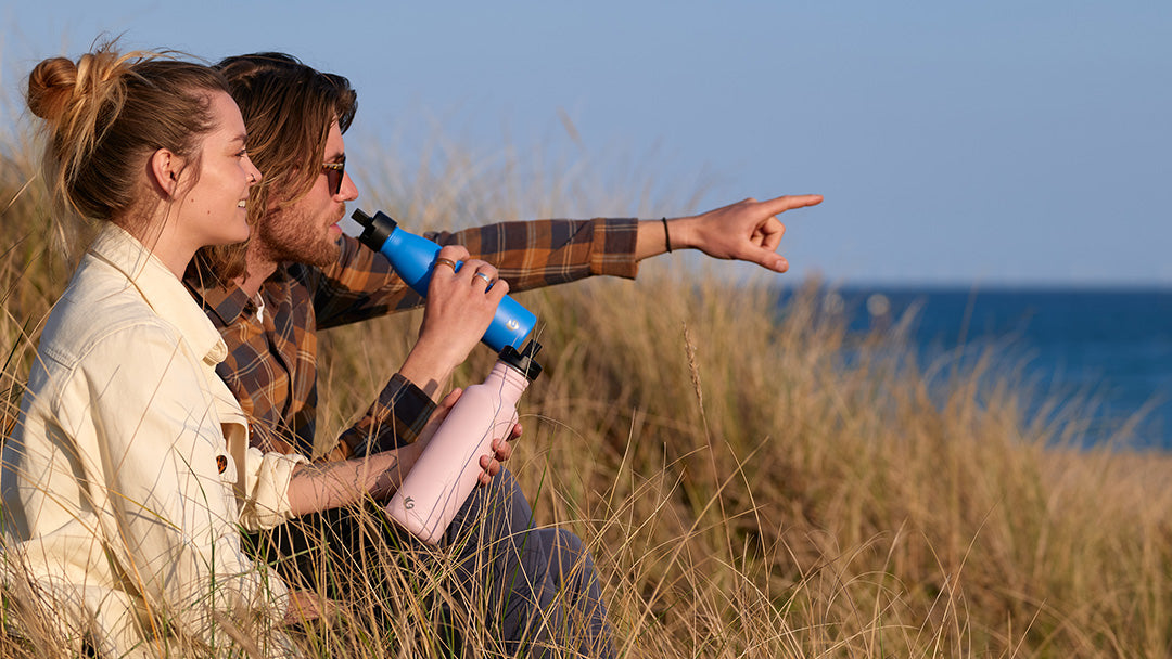 A couple sat in long grass enjoying a sea view, with One Green Bottle water bottles in hand