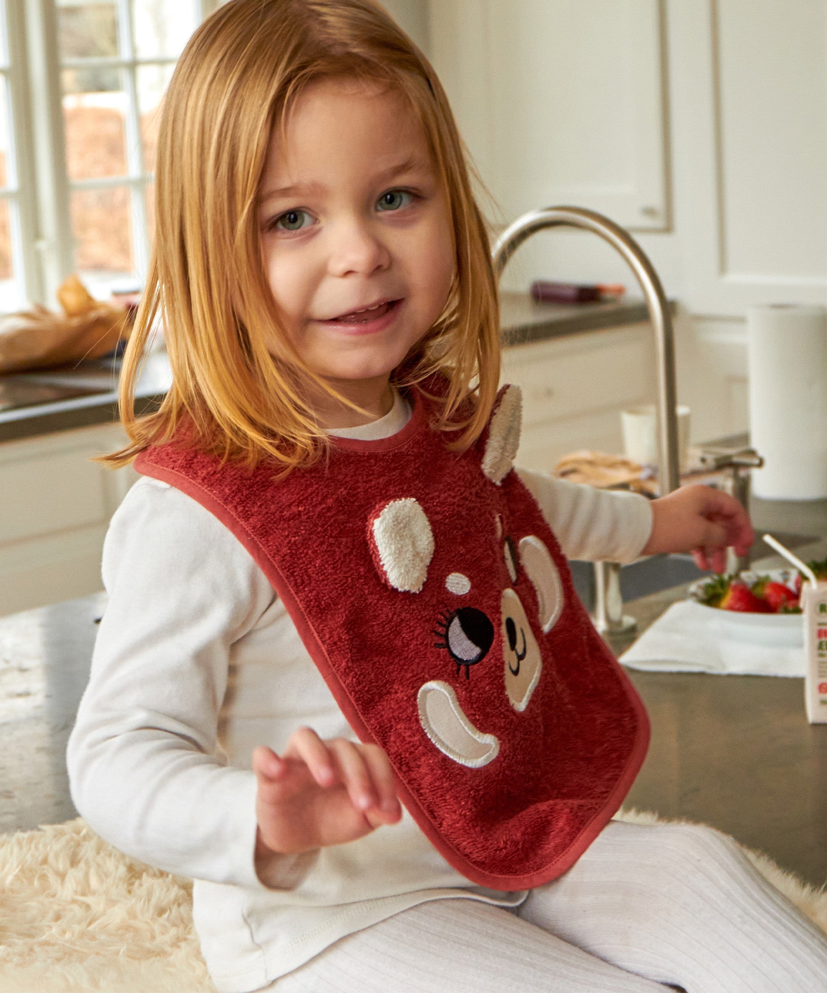 A child sat in a kitchen, wearing the Roommate Organic Cotton Baby Bib - Red Panda