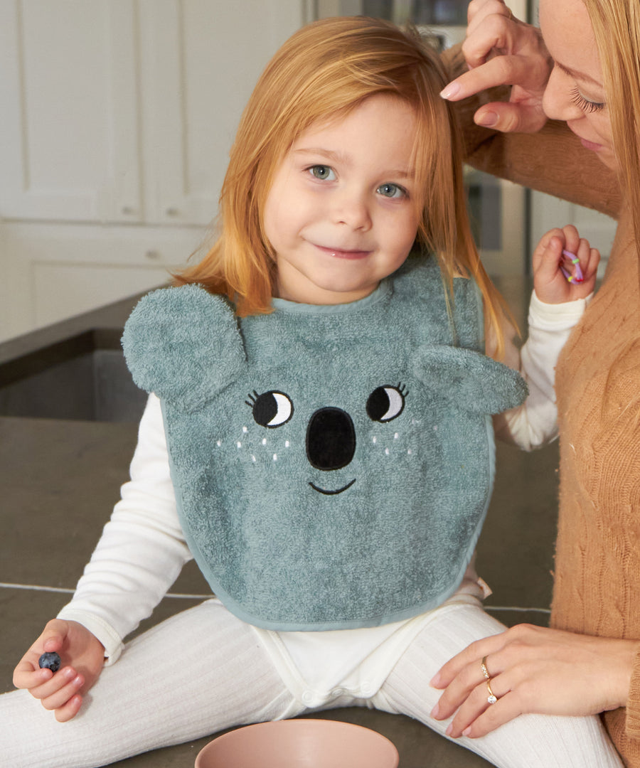 A child sat down with an adult, wearing the Roommate Organic Cotton Baby Bib - Koala