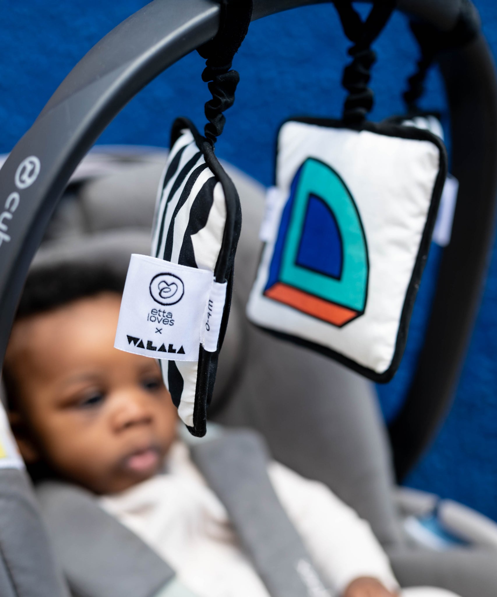 A baby sat in a car seat, looking at the Etta Loves x Camille Walala Sensory Hanging Squares which are hanging from the carry arm of the car seat