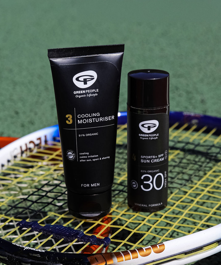 The Green People Sports+ Sun Cream and the Green People Cooling Moisturiser are both stood on top of a tennis racket, with a green playing field in the background