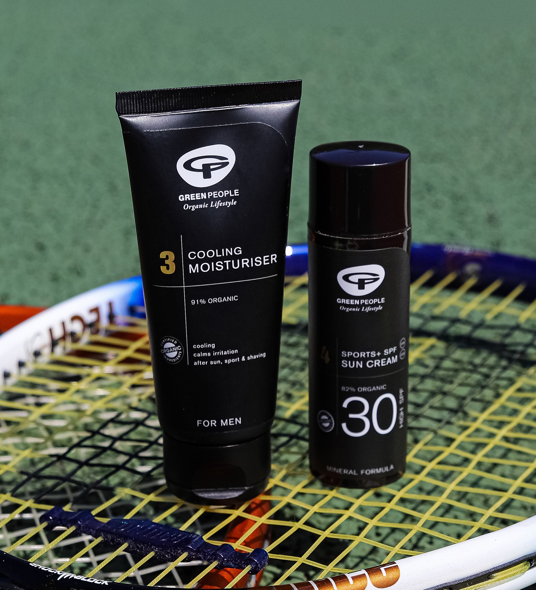 The Green People Sports+ Sun Cream and the Green People Cooling Moisturiser are both stood on top of a tennis racket, with a green playing field in the background