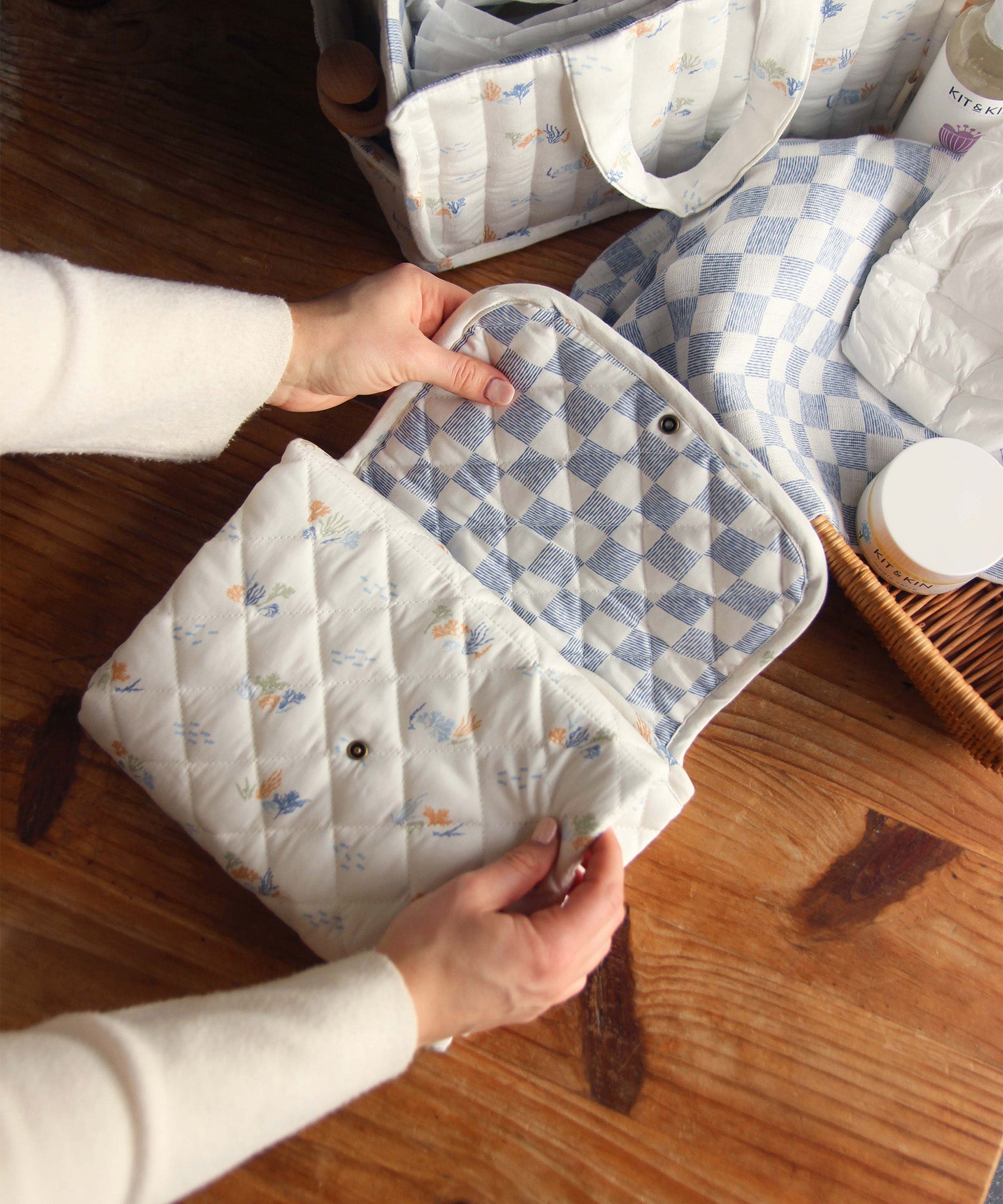 Avery Row Travel Changing Mat - Coastline in adults hands, un-poppered to show the blue and white checkerboard print inside on a wooden table.