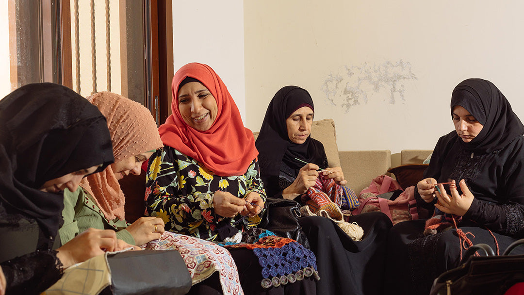 Five women sit together indoors, engaged in crafting. They are Syrian refugees, working on handmade items as part of the Oshana social enterprise, which empowers them to rebuild their lives.