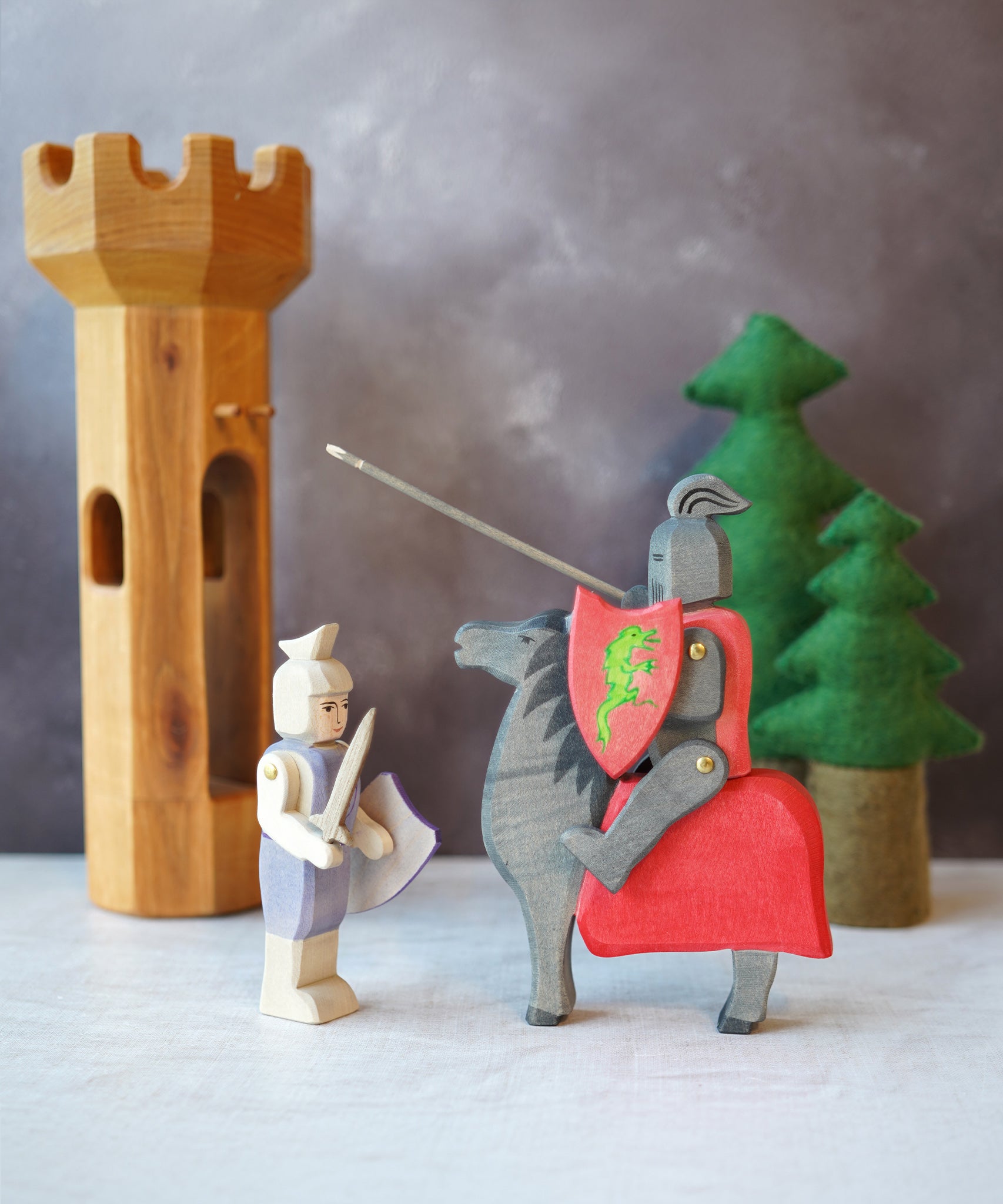 The Ostheimer Wooden Blue Standing Knight Figure standing next to the Black Knight and Horse set with trees and a wooden tower in the background.