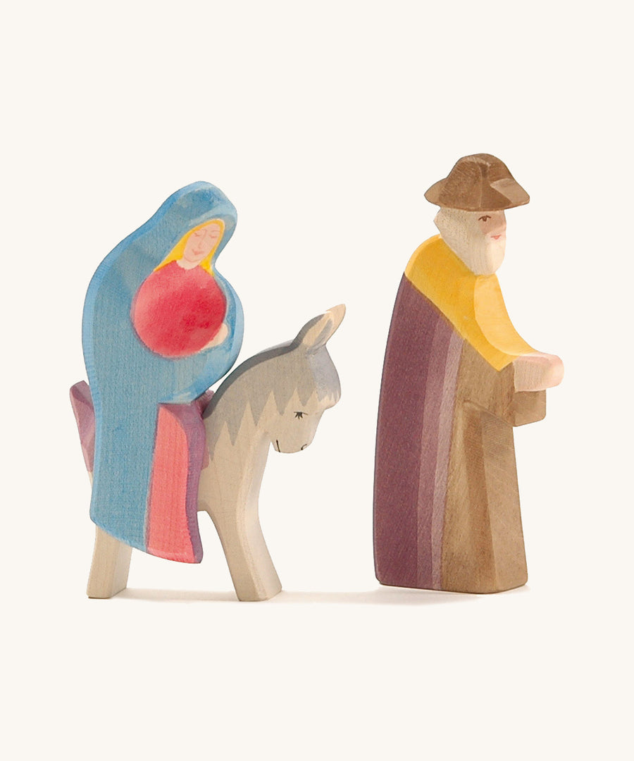 A wooden Mary Ostheimer figure wearing a red gown and blue cape sitting on a grey donkey with a Ostheimer wooden Joseph figure standing next to them on a cream background.