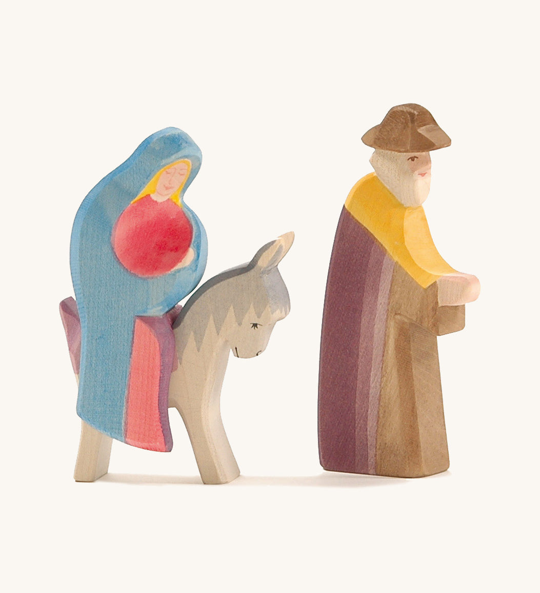 A wooden Mary Ostheimer figure wearing a red gown and blue cape sitting on a grey donkey with a Ostheimer wooden Joseph figure standing next to them on a cream background.