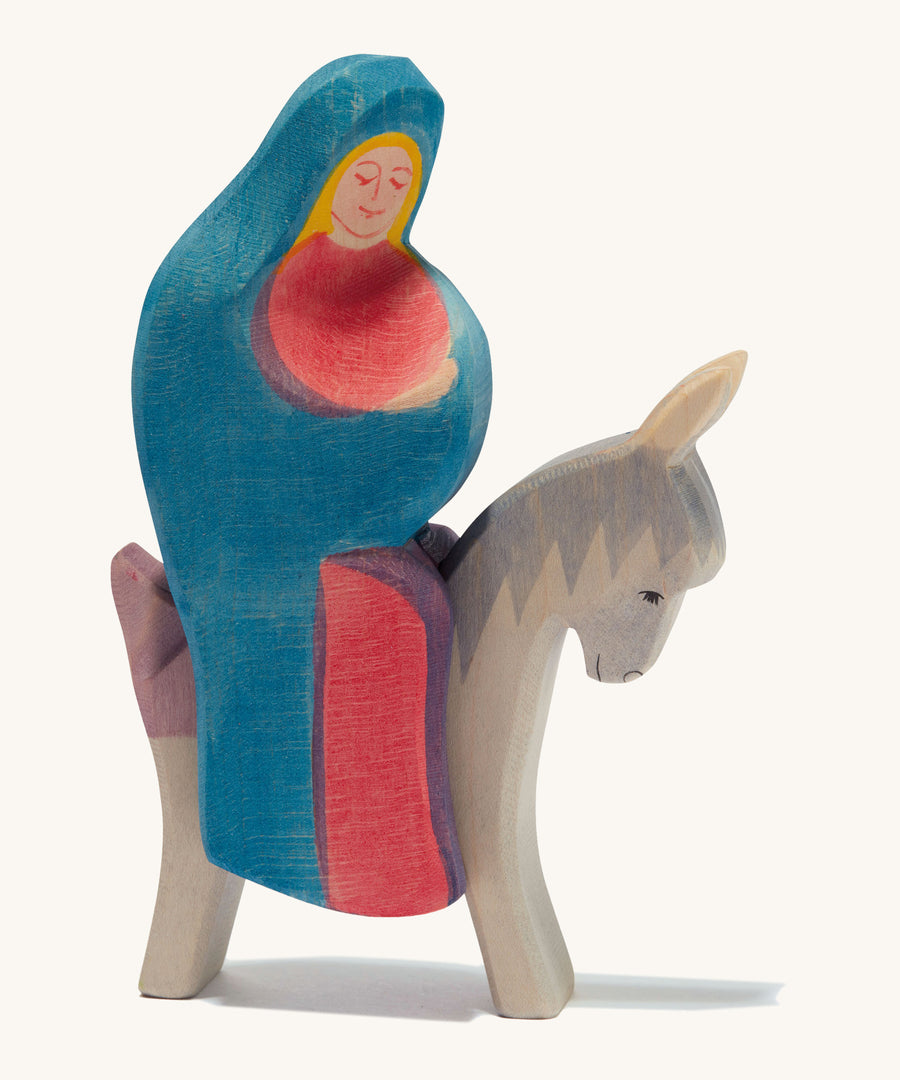 A wooden Mary Ostheimer figure wearing a red gown and blue cape sitting on a grey donkey on a cream background.