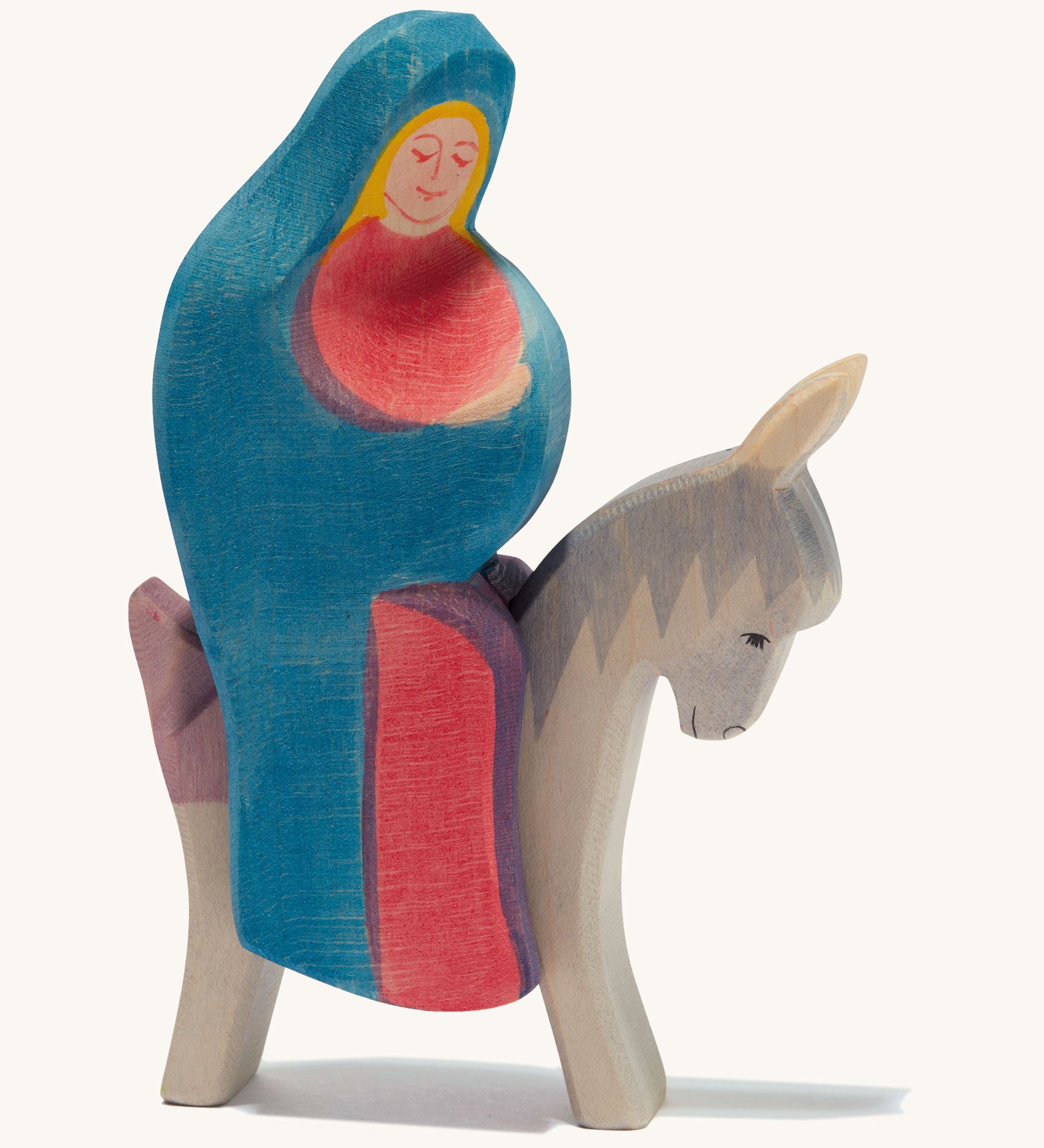 A wooden Mary Ostheimer figure wearing a red gown and blue cape sitting on a grey donkey on a cream background.