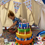 Bunting made with card and paper tape hung around an open tipi tent. There is a Babipur playmat inside the tent and a toy cake made by stacking a Grimm's rainbow and semi circles.