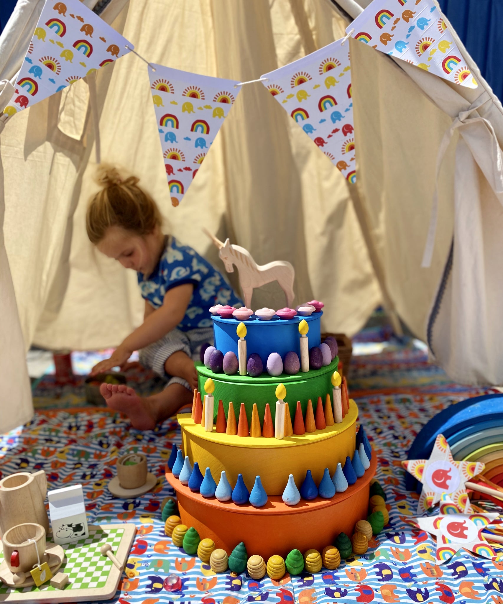 Bunting made with card and paper tape hung around an open tipi tent. There is a Babipur playmat inside the tent and a toy cake made by stacking a Grimm's rainbow and semi circles.