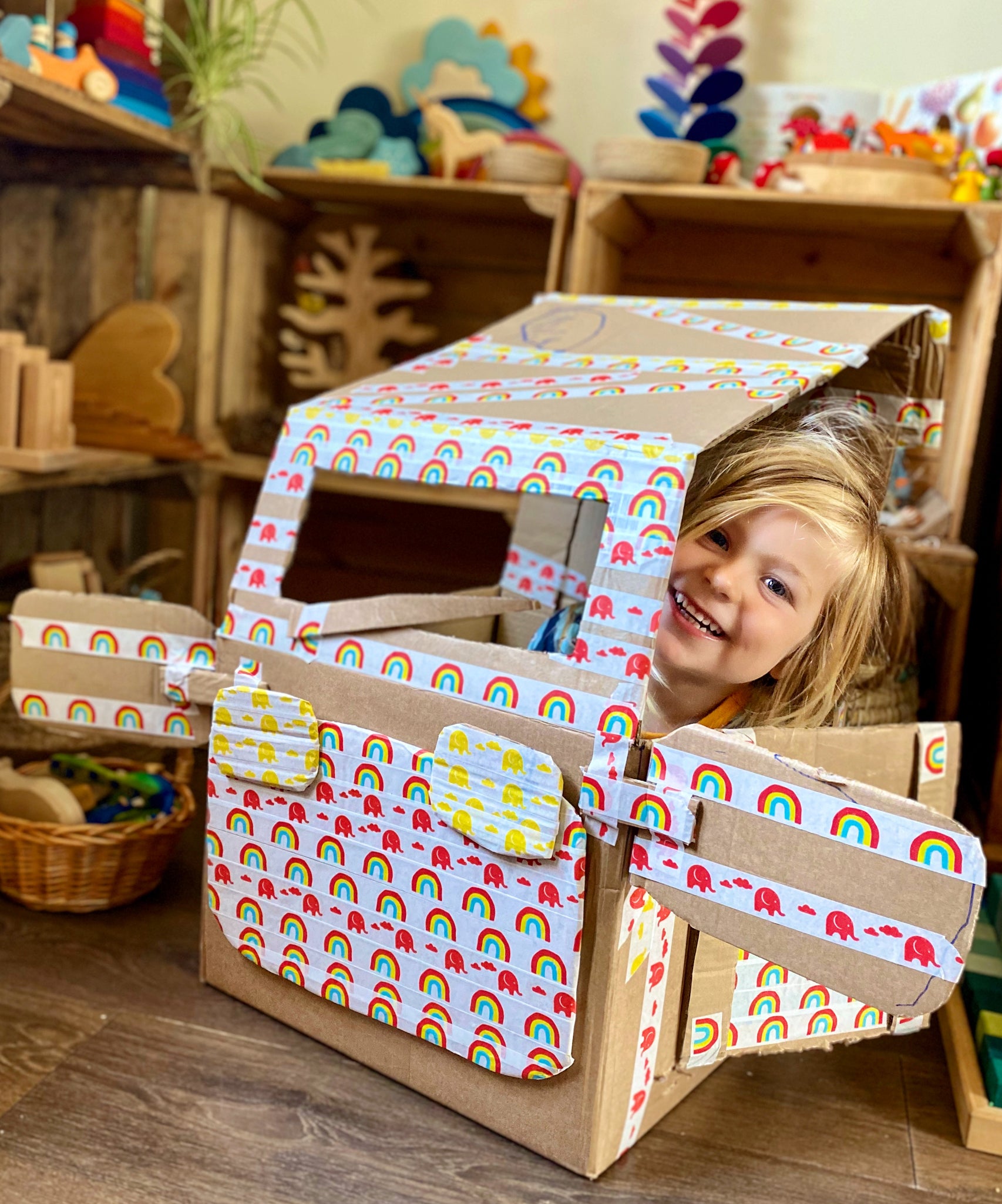 Child sitting in a car made out of cardboard. The car is decorated in Babipur paper tape. The photo has been taken in a playroom.