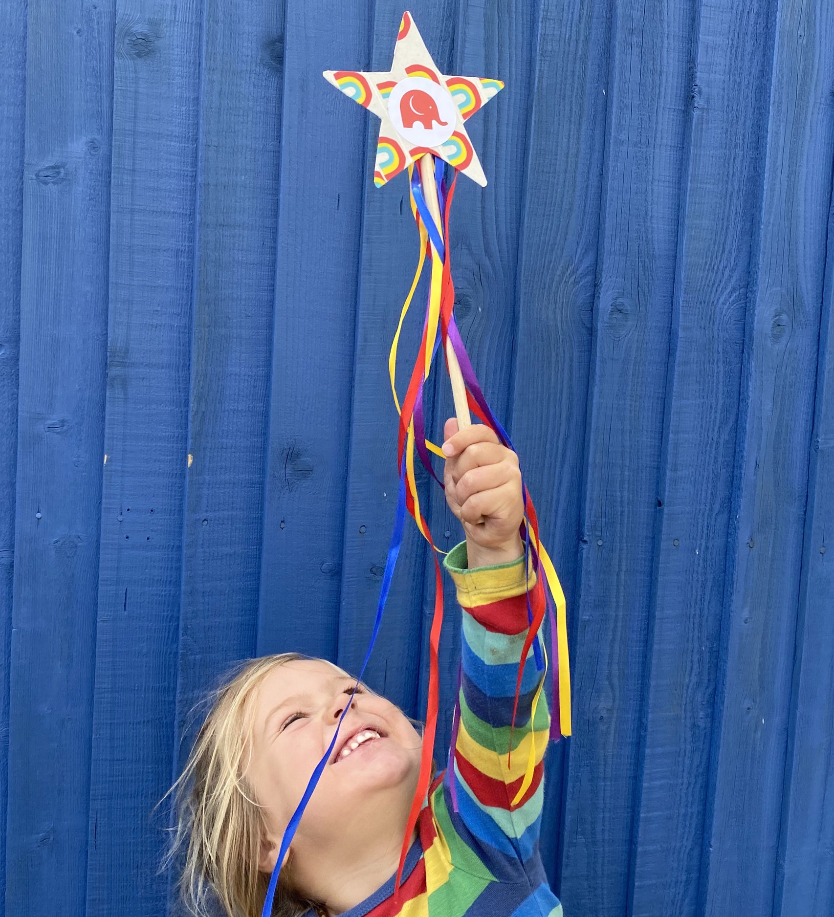 Child holding a cardboard star wand that's edged with Babipur rainbow tape. The child is stood in front of a blue shed.