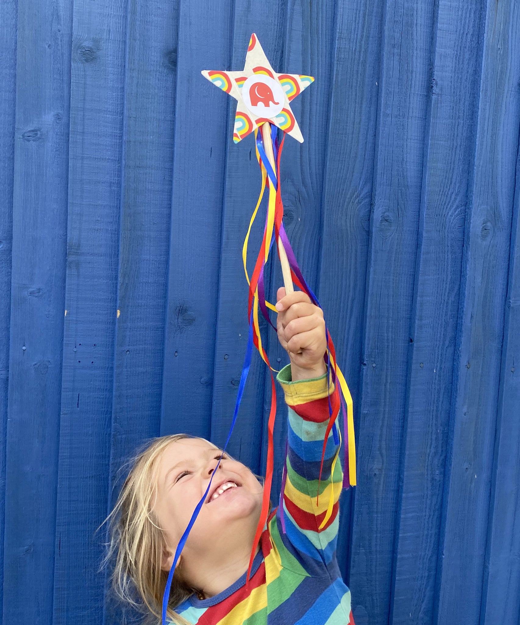 Child holding a cardboard star wand that's edged with Babipur rainbow tape. The child is stood in front of a blue shed.